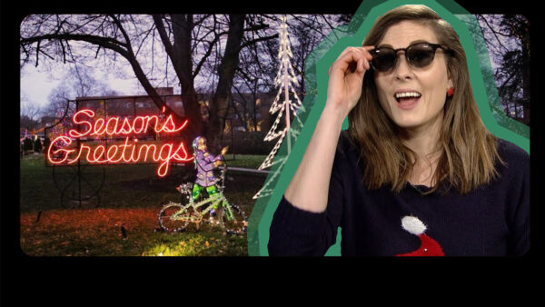 A woman in sunglasses and a sweater stands beside a "Merry Christmas" sign, smiling in a festive setting.