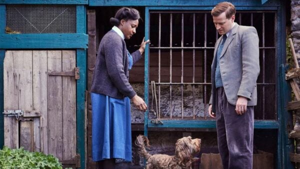 A man and woman stand in front of a home, looking at a dog between them