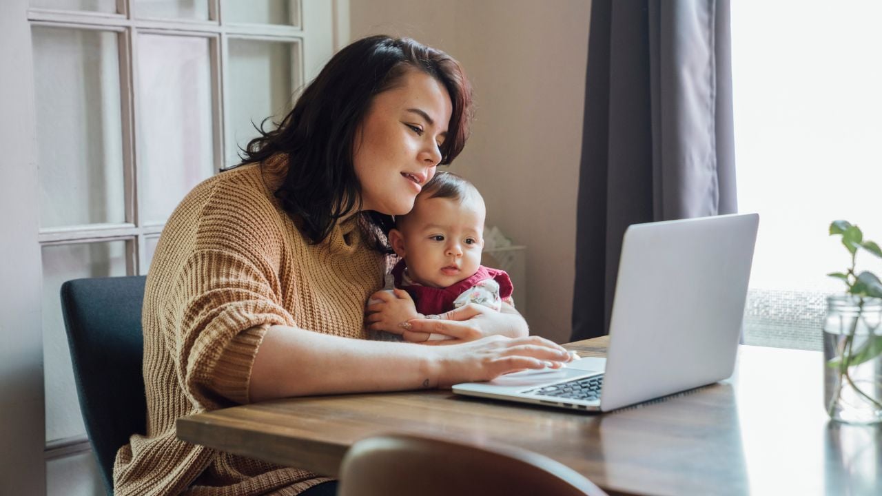 A woman sits at a table, holding a baby and looking at a computer