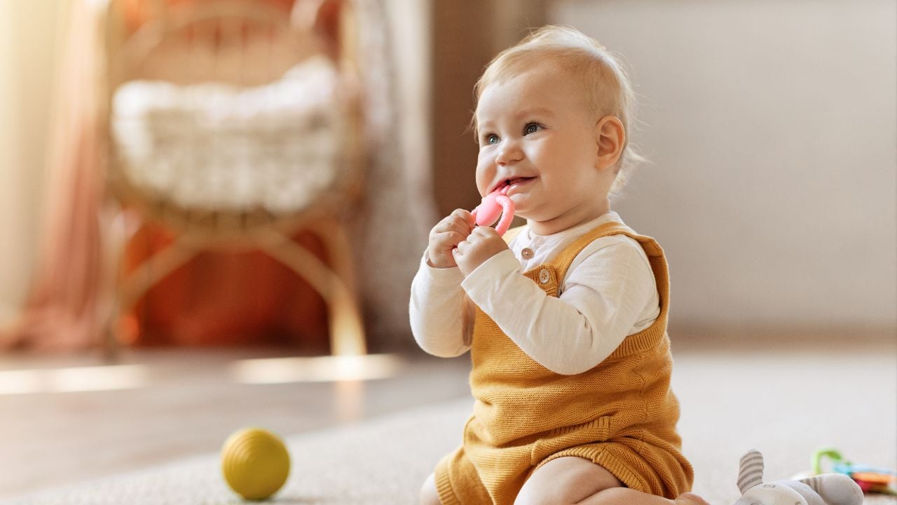A toddler sits on the floor chewing on a teething toy