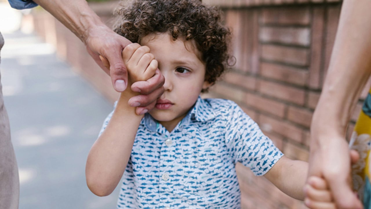 A young boy hides his face and tries not to cry while holding hands with an adult